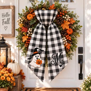 May include: A decorative wreath featuring green foliage, orange maple leaves, pinecones, and small white berries. A large black and white buffalo check fabric bow hangs from the centre, displaying a ghost with a pumpkin and the letter M.