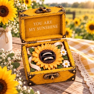 May include: A yellow wooden ring box with a hinged lid featuring the text YOU ARE MY SUNSHINE. The interior contains a large sunflower ornament with two ring slots holding a diamond band and a plain metal band, accented by small white flowers.