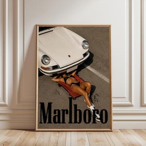 Peut inclure: Une affiche encadrée représentant une voiture de sport vintage blanche garée sur de l'asphalte. Une personne en bikini noir est allongée sur un chariot de mécanicien rouge sous le pare-chocs avant. Le mot Marlboro est écrit en noir en bas.