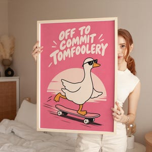 May include: A framed rectangular wall art print featuring a white goose wearing black sunglasses while riding a skateboard. The background is a vibrant pink with a light peach sun circle. The text reads OFF TO COMMIT TOMFOOLERY in white letters.