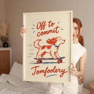 May include: A framed rectangular print on a cream background featuring a red and white dog riding a blue skateboard with red wheels. The text reads Off to commit at the top and Tomfoolery at the bottom in red script. The frame is light wood.