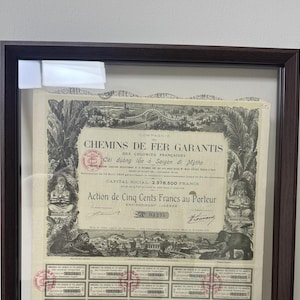 May include: A framed vintage stock certificate for the Compagnie des Chemins de Fer Garantis des Colonies Françaises. The document features intricate black and white illustrations of tropical landscapes, a tiger, and an elephant, with red stamps.