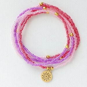 Puede incluir: Un brazalete de m&uacute;ltiples hilos hecho de cuentas rosas y moradas con un dije de sol dorado.
