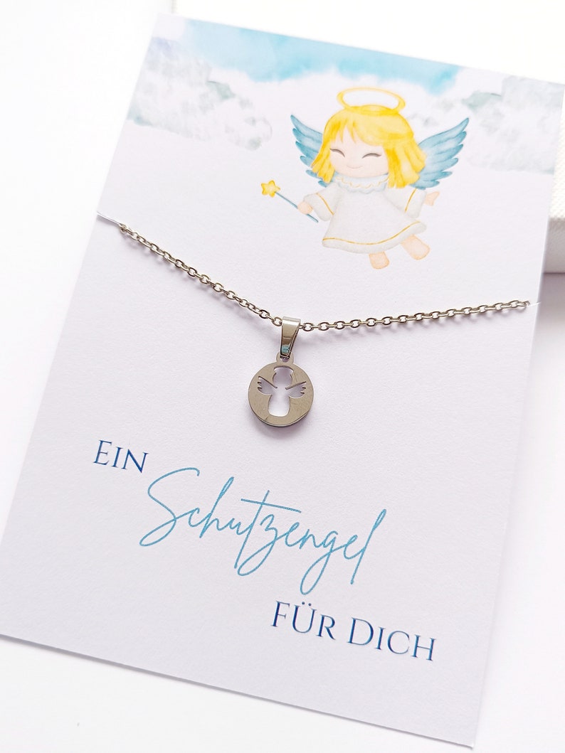 May include: A silver necklace with a pendant featuring a cutout angel design. The necklace is displayed on a white card with a watercolor illustration of an angel and the text "EIN Schutzengel F&Uuml;R DICH".