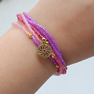 Puede incluir: Un brazalete de varias capas con cuentas rosas, moradas y blancas. El brazalete tiene un dije dorado en forma de sol.