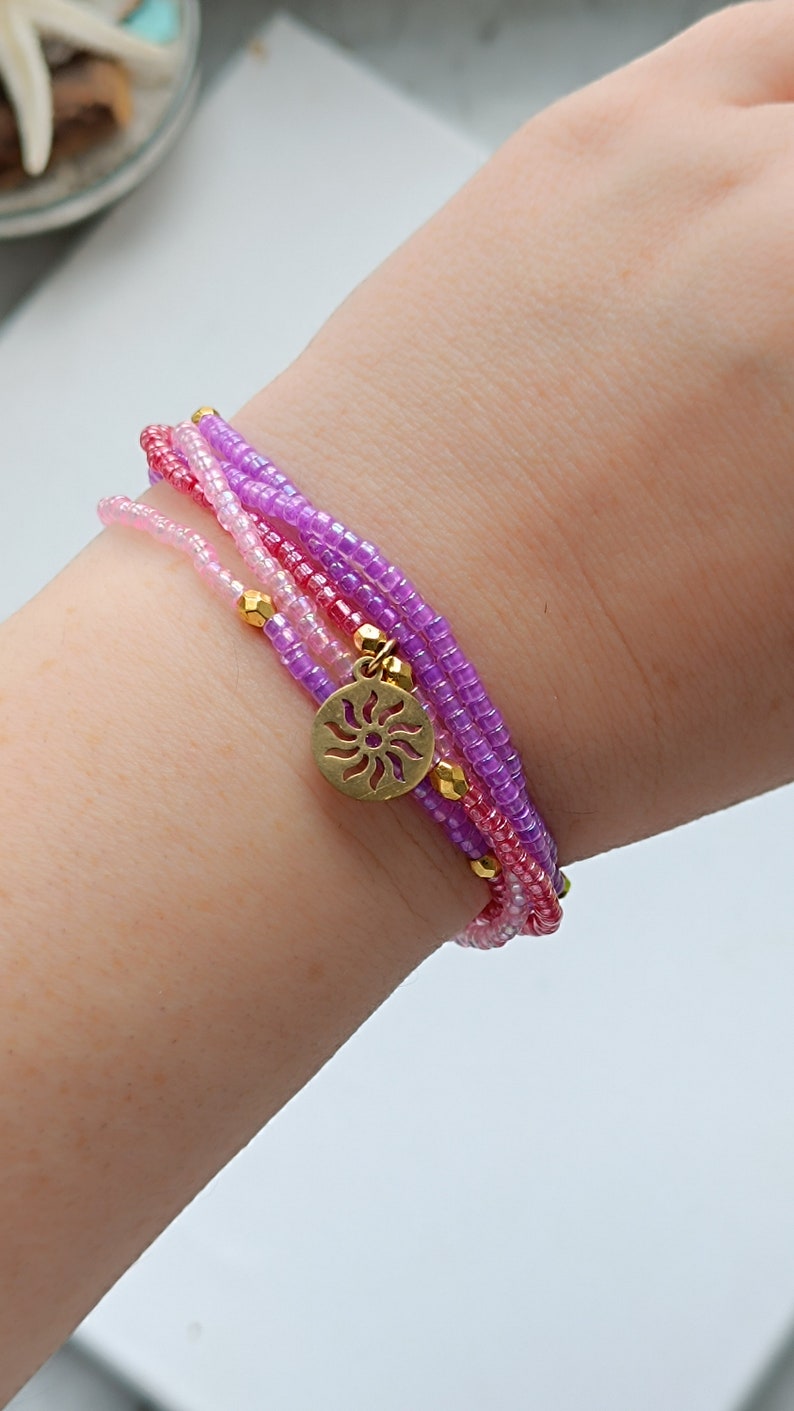 Puede incluir: Un brazalete de varias capas con cuentas rosas, moradas y blancas. El brazalete tiene un dije dorado en forma de sol.