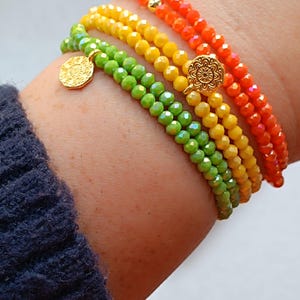 May include: A collection of beaded bracelets in a rainbow of colors: green, yellow, and orange. Each bracelet is made of small, faceted beads and features a gold-colored charm. The bracelets are stacked on a wrist.