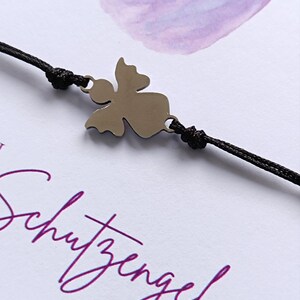 Peut inclure: Un bracelet avec un charm en forme d'ange argent&eacute; et un cordon noir. Le bracelet pr&eacute;sente un pendentif ange stylis&eacute; et est pos&eacute; sur un fond blanc avec le texte violet "Schutzengel F&Uuml;R DICH".