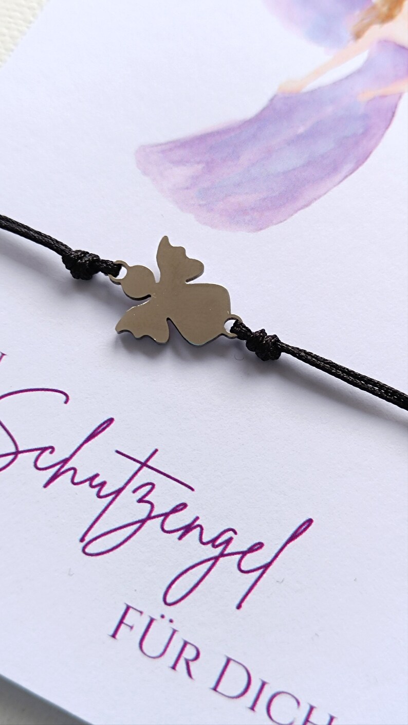 Peut inclure: Un bracelet avec un charm en forme d'ange argent&eacute; et un cordon noir. Le bracelet pr&eacute;sente un pendentif ange stylis&eacute; et est pos&eacute; sur un fond blanc avec le texte violet "Schutzengel F&Uuml;R DICH".