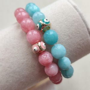 May include: Two stretch bracelets made of round polished beads. One bracelet features pink beads with a gold-tone metal charm containing pink enamel eyes. The second bracelet features light blue beads with a gold-tone metal charm containing teal enamel eyes.