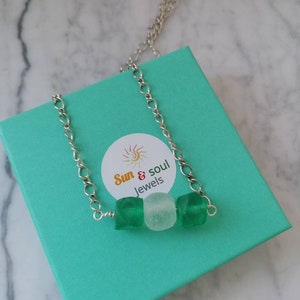 Peut inclure: Un collier en cha&icirc;ne argent&eacute;e avec trois perles&nbsp;: deux perles en verre vert et une perle en verre blanc. Le collier est dans une bo&icirc;te cadeau turquoise avec une &eacute;tiquette blanche qui dit&nbsp;&laquo;&nbsp;Sun & Soul Jewels&nbsp;&raquo;.