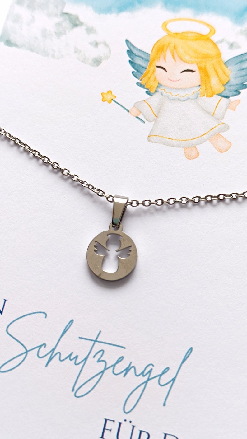 May include: A silver necklace with a round pendant featuring a cutout angel design. The pendant is hanging from a silver chain. The necklace is displayed on a white card with the text "Schutzengel f&uuml;r" in blue.