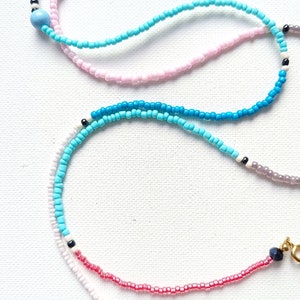 May include: A layered necklace with multiple strands of beads in various colors, including white, pink, blue, and black. The necklace features a mix of small round beads and larger round beads.