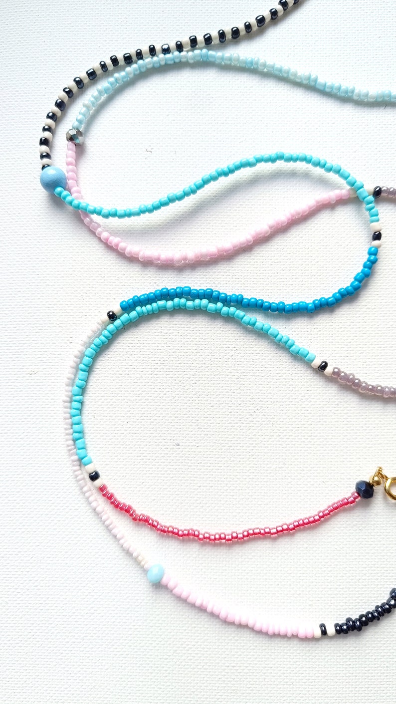 May include: A layered necklace with multiple strands of beads in various colors, including white, pink, blue, and black. The necklace features a mix of small round beads and larger round beads.