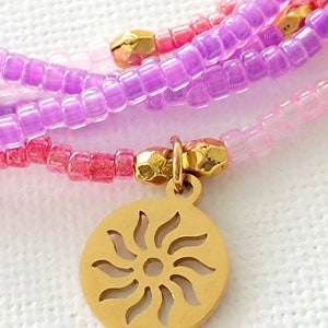 Puede incluir: Pulsera de m&uacute;ltiples hilos con cuentas rosas, moradas y blancas y un dije de sol dorado.