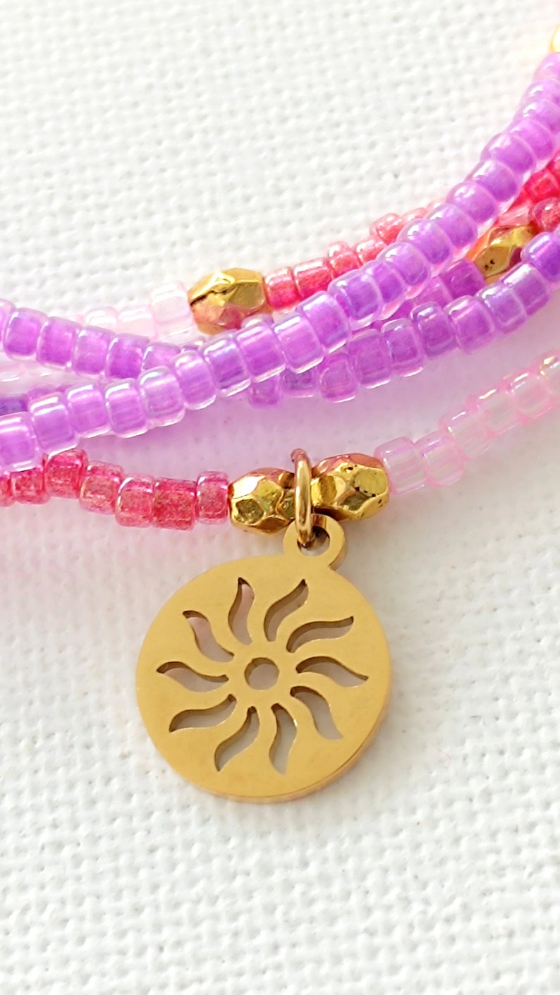 Puede incluir: Pulsera de m&uacute;ltiples hilos con cuentas rosas, moradas y blancas y un dije de sol dorado.
