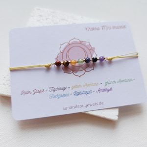 Peut inclure: Un bracelet Chakra Mini avec sept perles de pierres précieuses colorées, enfilées sur un cordon jaune. Le bracelet est présenté sur une carte blanche avec du texte et un motif floral. La carte comprend également le texte "sunandsouljewels.de".