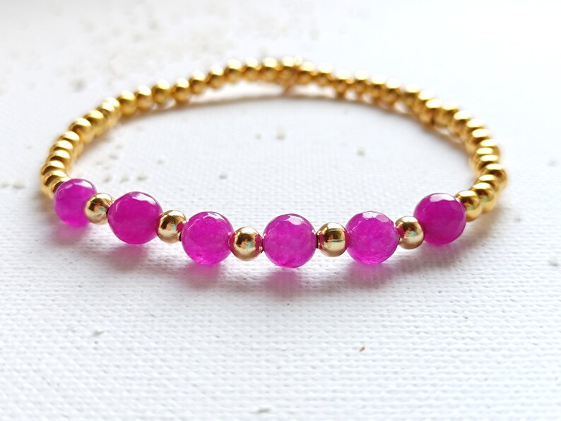 May include: A gold beaded bracelet with pink gemstone beads. The bracelet is made with small gold beads and larger pink beads.