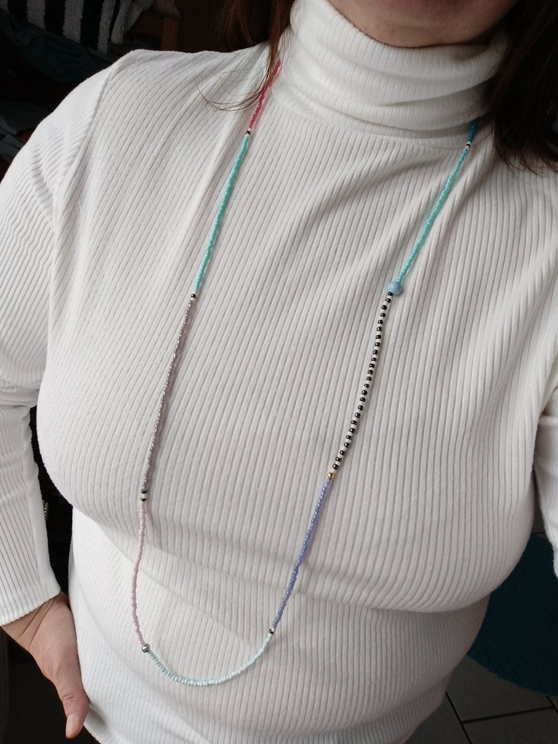 May include: A white turtleneck sweater with a multi-colored beaded necklace. The necklace has pink, blue, and black beads.