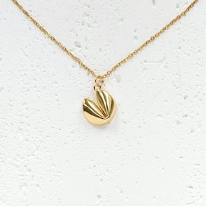 Puede incluir: Un collar dorado con un colgante en forma de galleta de la fortuna. El colgante tiene una superficie texturizada y detallada y cuelga de una delicada cadena dorada. El collar se muestra sobre un fondo blanco.