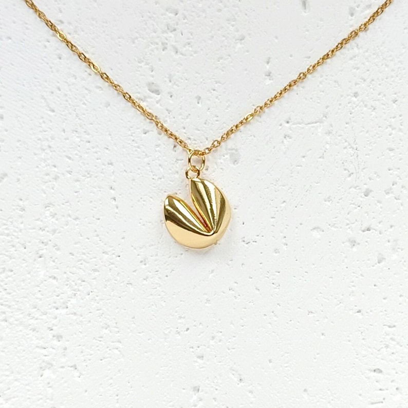 Puede incluir: Un collar dorado con un colgante en forma de galleta de la fortuna. El colgante tiene una superficie texturizada y detallada y cuelga de una delicada cadena dorada. El collar se muestra sobre un fondo blanco.