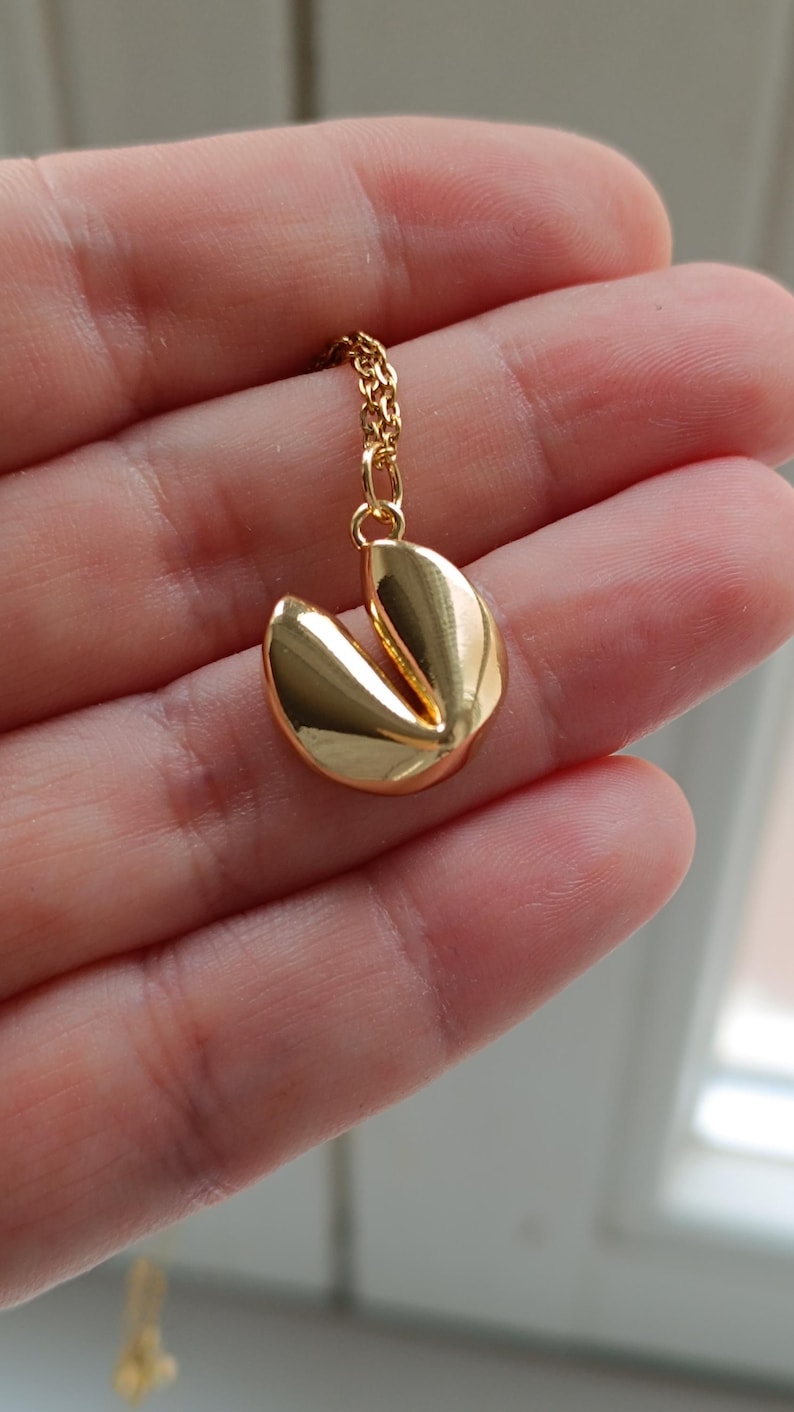 Puede incluir: Un collar con colgante de galleta de la fortuna dorada. El colgante es una r&eacute;plica detallada de una galleta de la fortuna, con una abertura en el medio. La cadena del collar tambi&eacute;n es dorada.