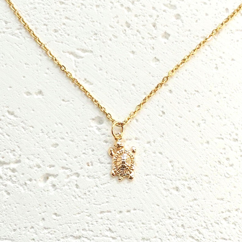 May include: A gold-colored necklace featuring a small turtle pendant. The turtle pendant is intricately detailed, and the necklace chain is delicate. The necklace is displayed against a textured white background.