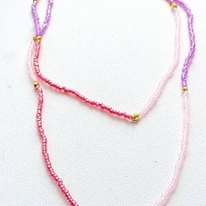 Puede incluir: Un collar de capas con cuentas rosas, moradas y rojas y un dije de sol dorado.