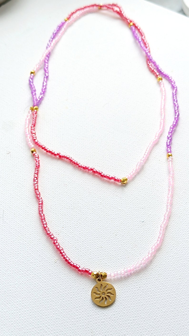 Puede incluir: Un collar de capas con cuentas rosas, moradas y rojas y un dije de sol dorado.