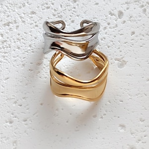 May include: Two wavy rings, one silver and one gold, stacked on top of each other. The rings are simple and modern in design.