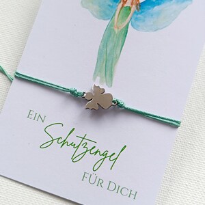 Peut inclure: Un bracelet avec un charm ange argent&eacute; et un cordon vert menthe est pr&eacute;sent&eacute; sur une carte avec une illustration d'ange &agrave; l'aquarelle. La carte porte l'inscription "EIN Schutzengel F&Uuml;R DICH" en vert.