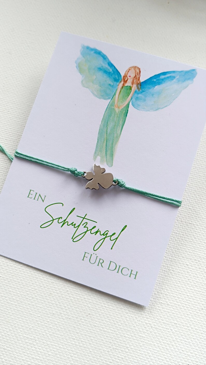 Peut inclure: Un bracelet avec un charm ange argent&eacute; et un cordon vert menthe est pr&eacute;sent&eacute; sur une carte avec une illustration d'ange &agrave; l'aquarelle. La carte porte l'inscription "EIN Schutzengel F&Uuml;R DICH" en vert.