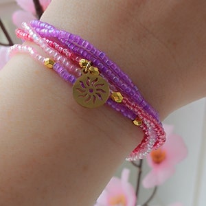 Puede incluir: Un brazalete de varias capas con cuentas rosas, moradas y blancas. Un dije dorado con un dise&ntilde;o de sol cuelga del brazalete.