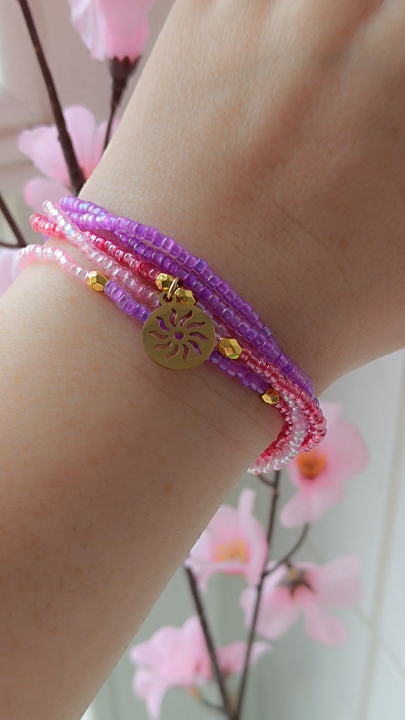 Puede incluir: Un brazalete de varias capas con cuentas rosas, moradas y blancas. Un dije dorado con un dise&ntilde;o de sol cuelga del brazalete.