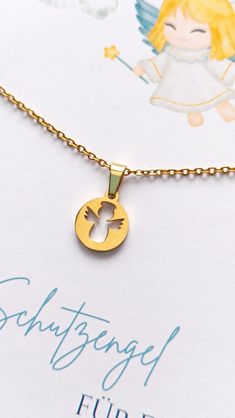 May include: A gold chain necklace with a round pendant featuring a cutout angel design. The pendant is on a white background with the text "Schutzengel f&uuml;r" in blue.