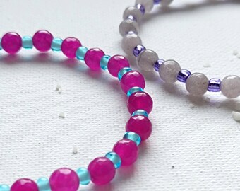 May include: Two beaded bracelets. One bracelet is made with pink and blue beads. The other bracelet is made with gray and purple beads.