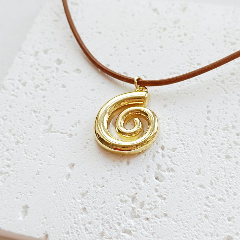 May include: A gold-coloured pendant in a spiral design hangs from a brown cord necklace. The pendant is a shiny, metallic gold colour. The necklace cord is a thin, smooth brown material. The background is a textured, off-white surface.