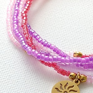 Puede incluir: Un brazalete de m&uacute;ltiples hilos hecho de peque&ntilde;as cuentas rosadas, moradas y blancas con un dije de sol dorado.