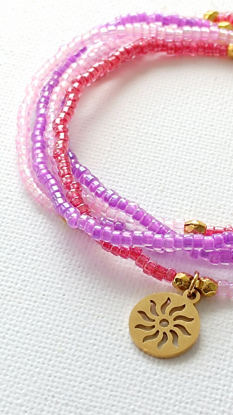 Puede incluir: Un brazalete de m&uacute;ltiples hilos hecho de peque&ntilde;as cuentas rosadas, moradas y blancas con un dije de sol dorado.