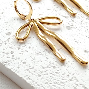 May include: Gold-toned bow-shaped earrings with a textured, wavy design. The earrings are dangling and have a hoop closure.