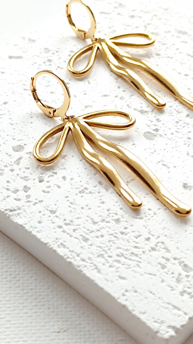 May include: Gold-toned bow-shaped earrings with a textured, wavy design. The earrings are dangling and have a hoop closure.