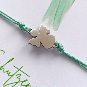 Peut inclure: Bracelet avec un charm en forme d'ange argent&eacute; et un cordon vert menthe. Le charm est au centre du bracelet. Le bracelet est pos&eacute; sur une carte blanche avec du texte vert et une aquarelle.