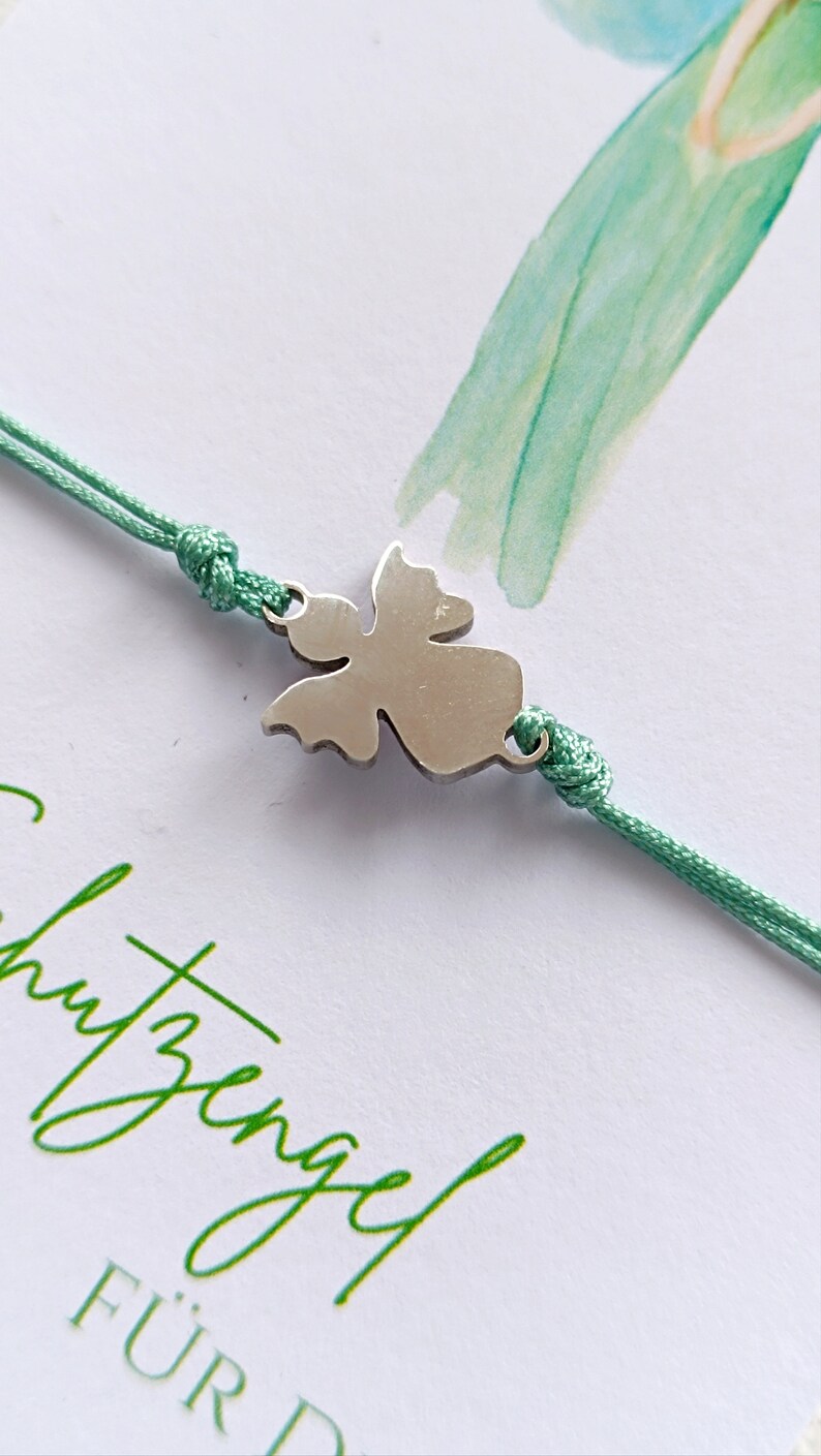 Peut inclure: Bracelet avec un charm en forme d'ange argent&eacute; et un cordon vert menthe. Le charm est au centre du bracelet. Le bracelet est pos&eacute; sur une carte blanche avec du texte vert et une aquarelle.