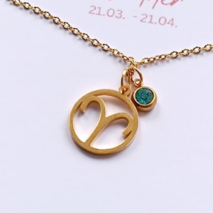 May include: Gold-colored necklace with a pendant featuring the Aries zodiac symbol and a small teal gemstone. The necklace is on a white card with the word "Widder" and the dates 21.03. - 21.04. printed in pink.