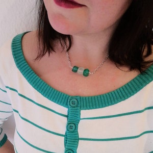 Peut inclure: Un collier en cha&icirc;ne argent avec trois perles en verre vert et blanc. Le collier est port&eacute; par une personne aux cheveux fonc&eacute;s et portant une chemise &agrave; rayures vertes et blanches.