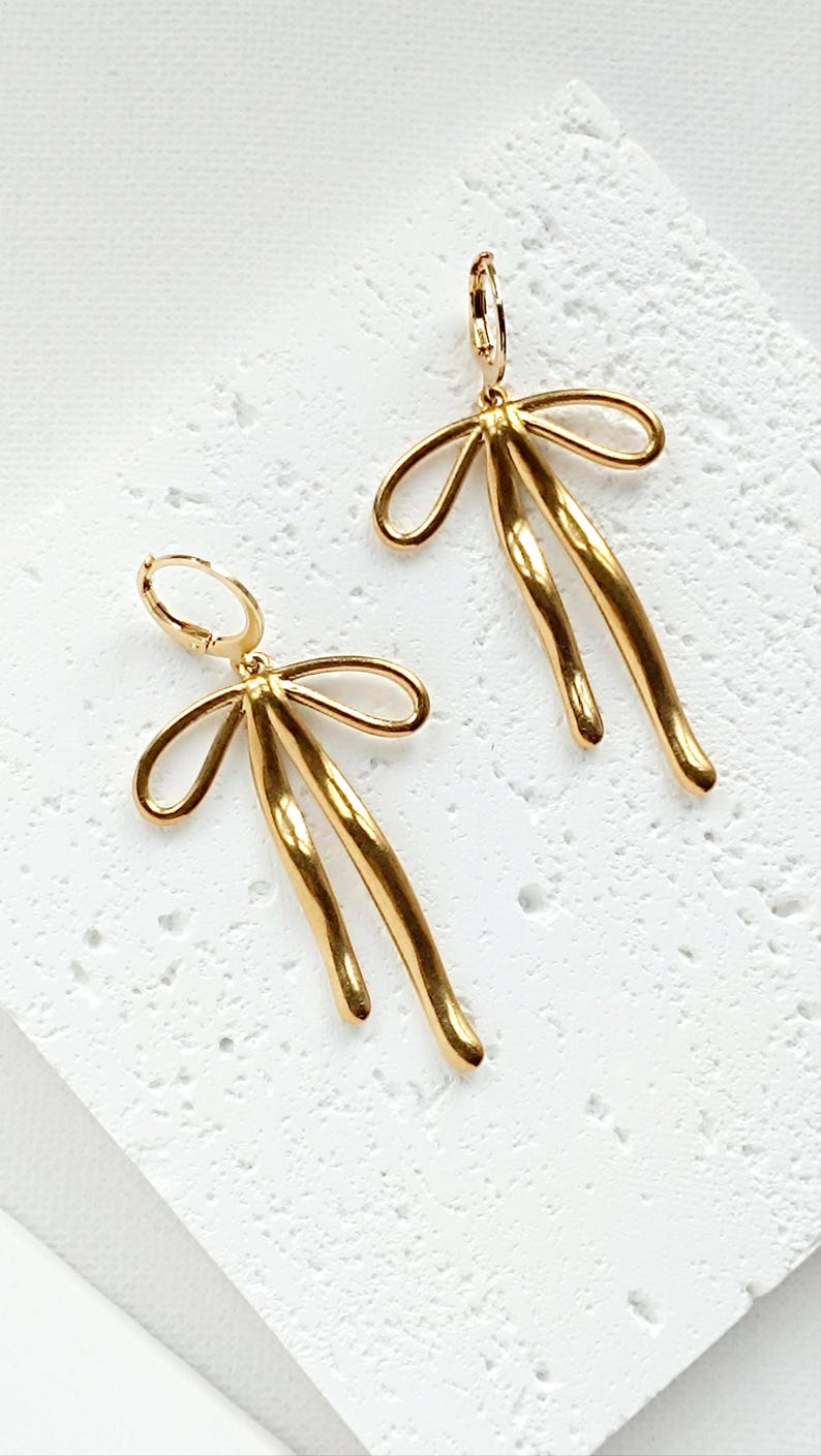 May include: A pair of gold-toned earrings with a bow design. The earrings are made of metal and have a hoop closure.