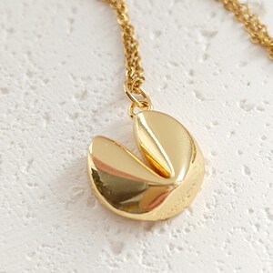 Puede incluir: Un collar con colgante de galleta de la fortuna de color dorado. El colgante es una r&eacute;plica detallada de una galleta de la fortuna, con un dise&ntilde;o dividido. La cadena del collar tambi&eacute;n es dorada y delicada. El collar se exhibe sobre una superficie blanca.
