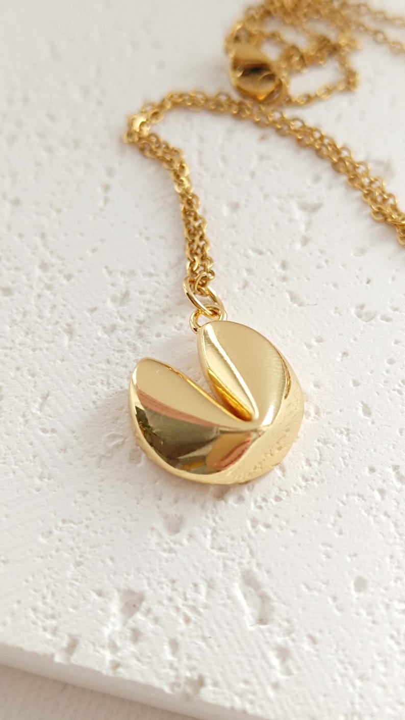 Puede incluir: Un collar con colgante de galleta de la fortuna de color dorado. El colgante es una r&eacute;plica detallada de una galleta de la fortuna, con un dise&ntilde;o dividido. La cadena del collar tambi&eacute;n es dorada y delicada. El collar se exhibe sobre una superficie blanca.