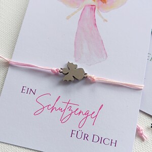 Peut inclure: Bracelet en fil rose avec un charm en forme d'ange argent&eacute;, pr&eacute;sent&eacute; sur une carte avec une illustration d'ange. La carte porte le texte allemand "EIN Schutzengel F&Uuml;R DICH" en &eacute;criture rose.
