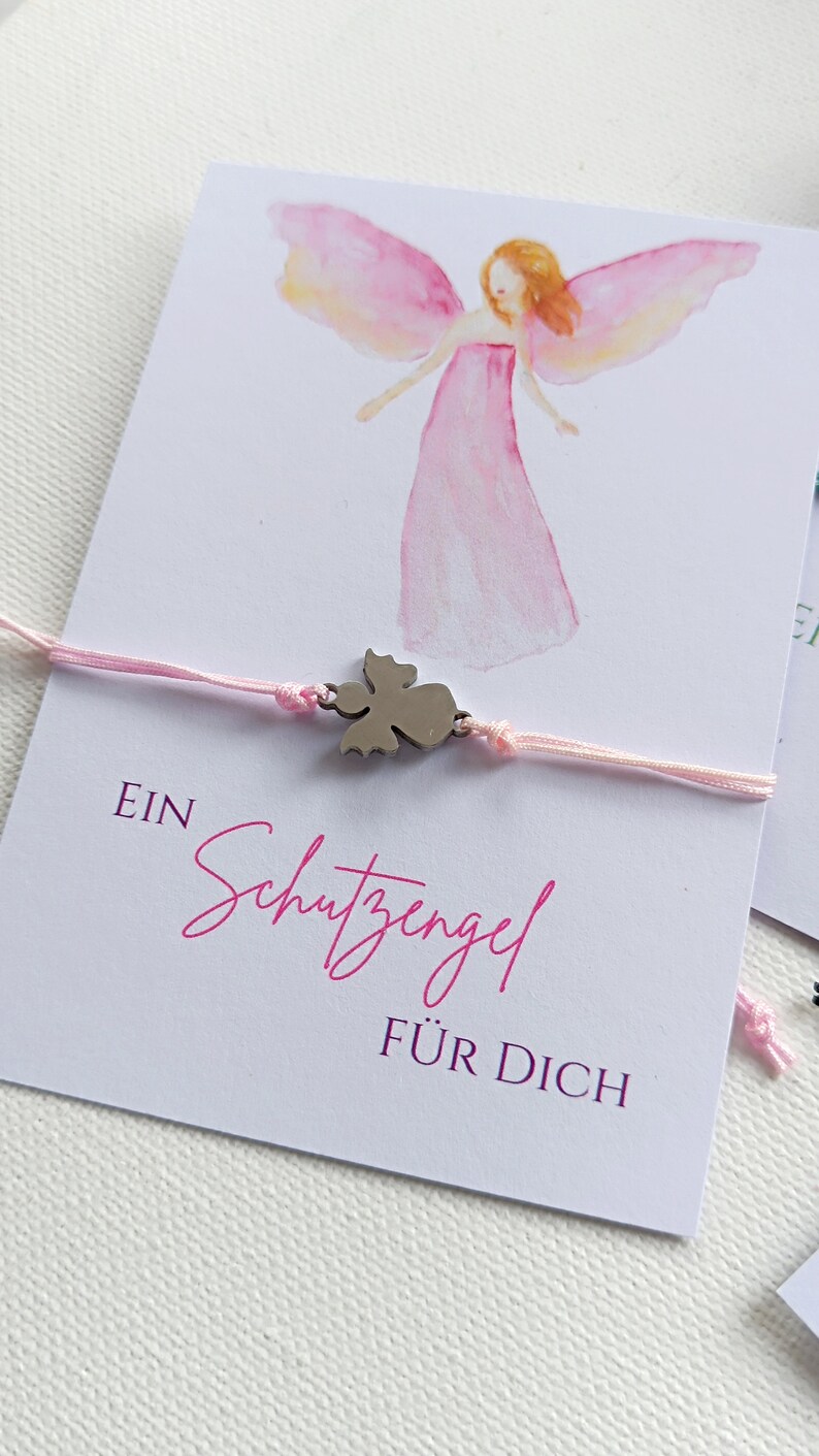Peut inclure: Bracelet en fil rose avec un charm en forme d'ange argent&eacute;, pr&eacute;sent&eacute; sur une carte avec une illustration d'ange. La carte porte le texte allemand "EIN Schutzengel F&Uuml;R DICH" en &eacute;criture rose.